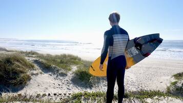 Eine Person hält ein Surfbrett am Strand, im Hintergrund Dünen und Meer.