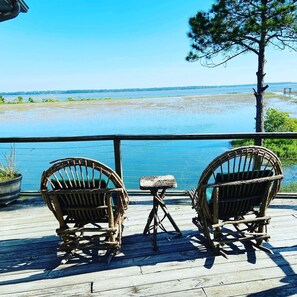 A wooden deck with two wicker chairs and a small table, overlooking a calm body of water and distant trees.