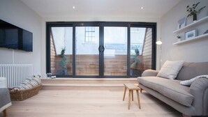 Una sala de estar moderna con un amplio ventanal corredera que da a una terraza de madera, un sofá gris, un taburete de madera y un banco de mimbre.