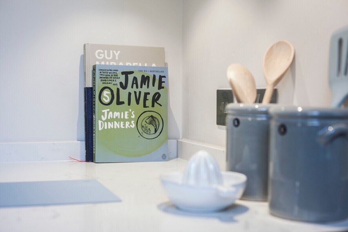 A kitchen counter with a book titled 'Jamie Oliver's Dinners' by Guy Malizia, wooden spoons, a juicer, and two grey mugs.