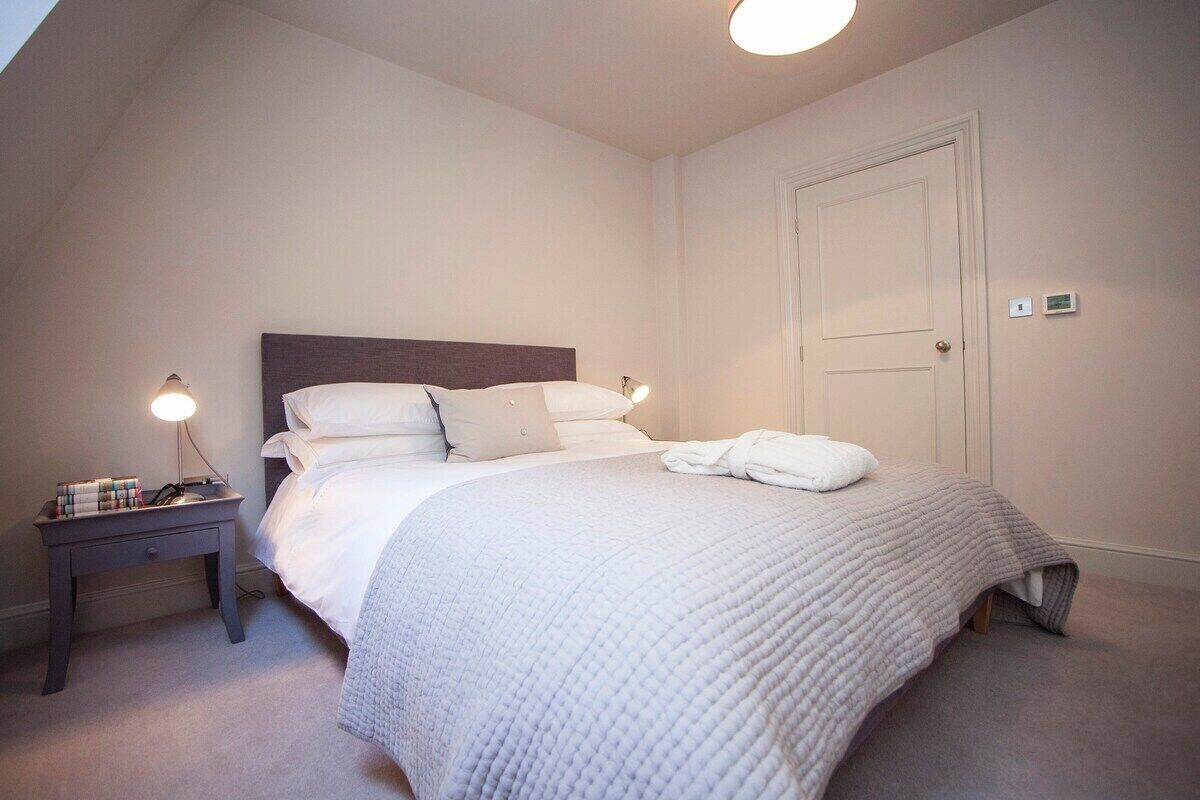 A neatly made bed with a white duvet and pillows, a bedside table with a lamp, and a door with a light switch.