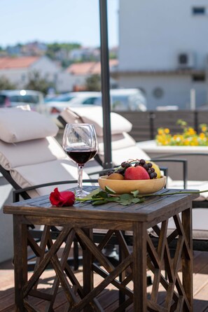 Una mesa de madera con una copa de vino tinto, un cuenco de fruta y una rosa roja en una terraza con sillones y vistas a edificios al fondo.
