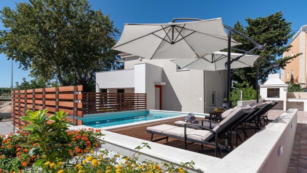 A modern outdoor pool area with lounge chairs and umbrellas, surrounded by a wooden fence and flowering plants. - Apartment house Ivan John (Podstrana)