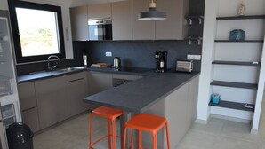 A modern kitchen with a large island, two orange stools, stainless steel appliances, and a window providing natural light. - Comfortable 3-bedroom villa 500 metres from LOZARI beach (Palasca)