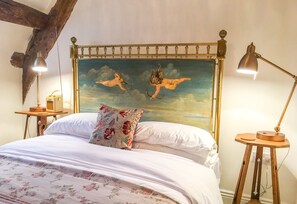 Quarto com cama grande, colcha floral, travesseiro decorativo, pintura de querubins no teto e duas luminárias de cabeceira.