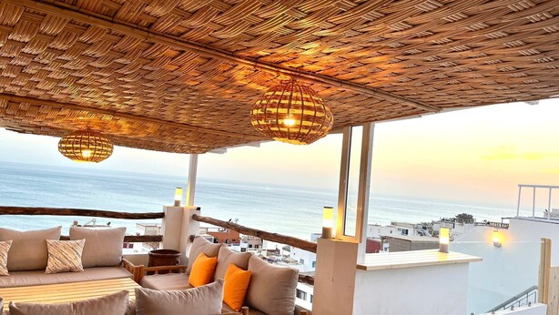 A balcony with a woven ceiling, cushioned seating, and a view of the sea. - BigBlue Hostel Surf & Coworking (Taghazout)