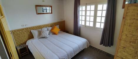 Un dormitorio con cama, cabecera, ventana con cortinas, espejo montado en la pared y una puerta.