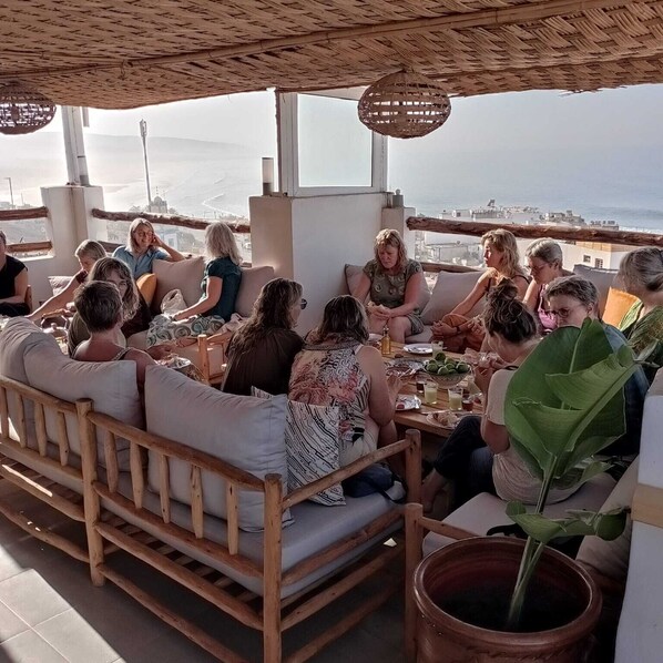 A group of people seated on wooden furniture under a thatched roof, with a view of the sea and buildings in the background. - BigBlue Hostel Surf & Coworking (Taghazout)