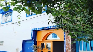 A building with a blue and white facade, a prominent orange door, and a small potted plant in front. - BigBlue Hostel Surf & Coworking (Taghazout)
