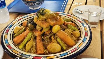 A dining table with a large plate of couscous, a bowl of fruit, and a bottle of water.