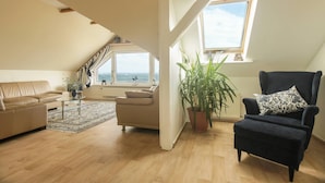 A spacious loft apartment with a high ceiling, wooden beams, a large window overlooking the sea, a black armchair, a beige sofa, and a potted plant. - Strandhaus Brunhild (Timmendorfer Strand)