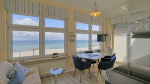 A room with a view of the beach, a dining area with chairs, a sofa with cushions, and a small table with a lamp. - Strandhaus Brunhild (Timmendorfer Strand)