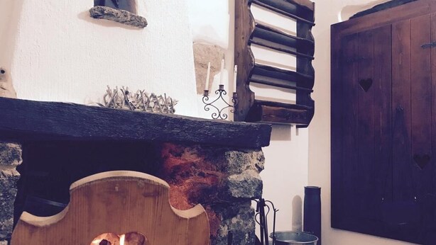 A rustic interior with a stone fireplace, a wooden bench with a heart-shaped cutout, a metal bucket, and a wooden door. - Cottage in Sardegna Barbagia (Fonni)