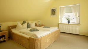 Une chambre avec un lit en osier, une table de chevet, une fenêtre avec des rideaux et un radiateur.