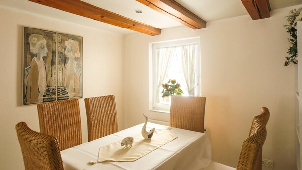 Un coin repas avec une table blanche, des chaises en osier, un tableau et une fenêtre avec des rideaux.