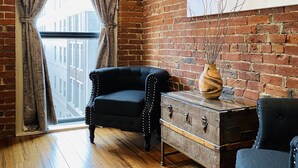 A room with a brick wall, a black upholstered sofa, a wooden chest of drawers, and a large window with curtains.