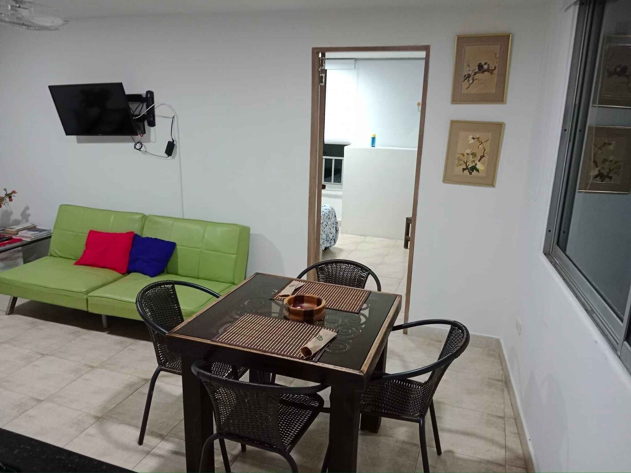 Une pièce avec un canapé vert, une petite table entourée de chaises, un téléviseur fixé au mur et des tableaux encadrés accrochés au mur.