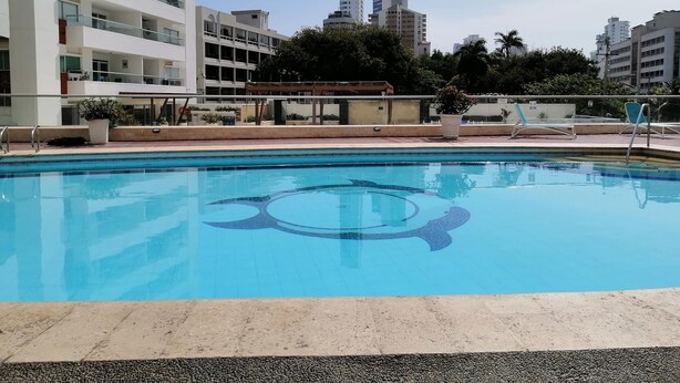Una piscina con diseño de agua azul cristalina, rodeada por una solana de concreto. Al fondo, edificios de varios pisos con balcones y un cielo despejado.
