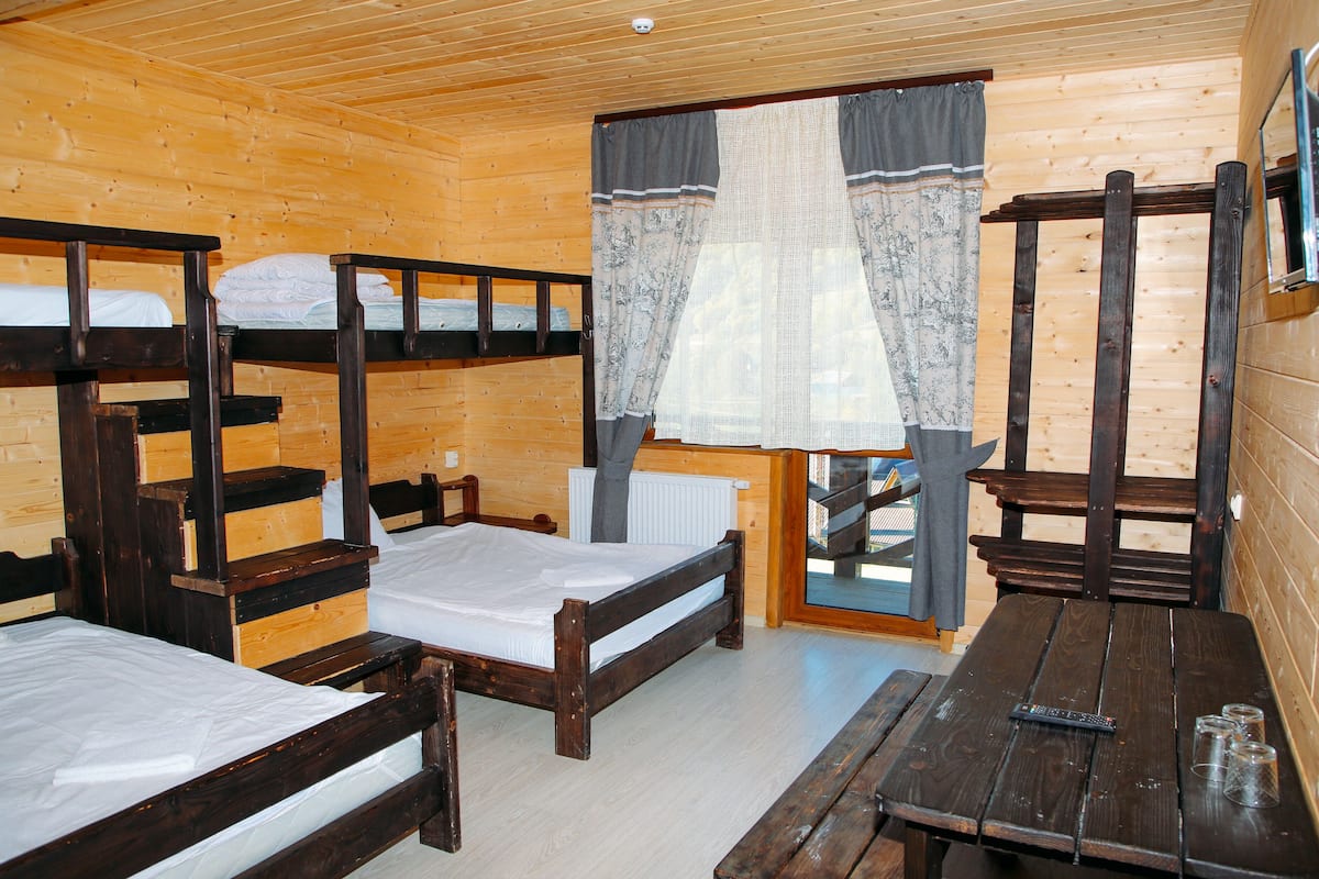 A cabin with wooden walls and floors, featuring bunk beds, a wooden table, and a window with curtains.