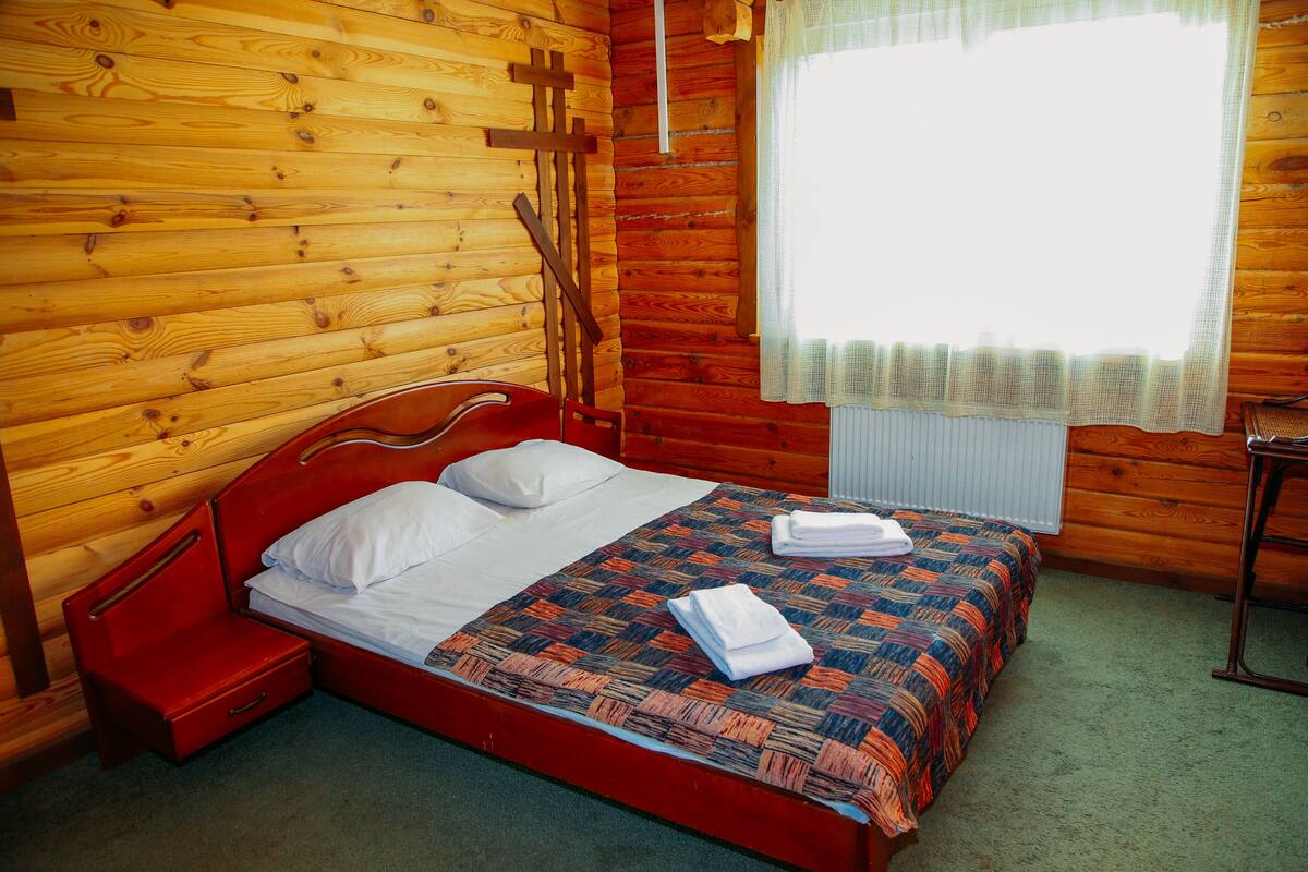 A wooden cabin bedroom with a single bed, a small nightstand, and a window with a sheer curtain.
