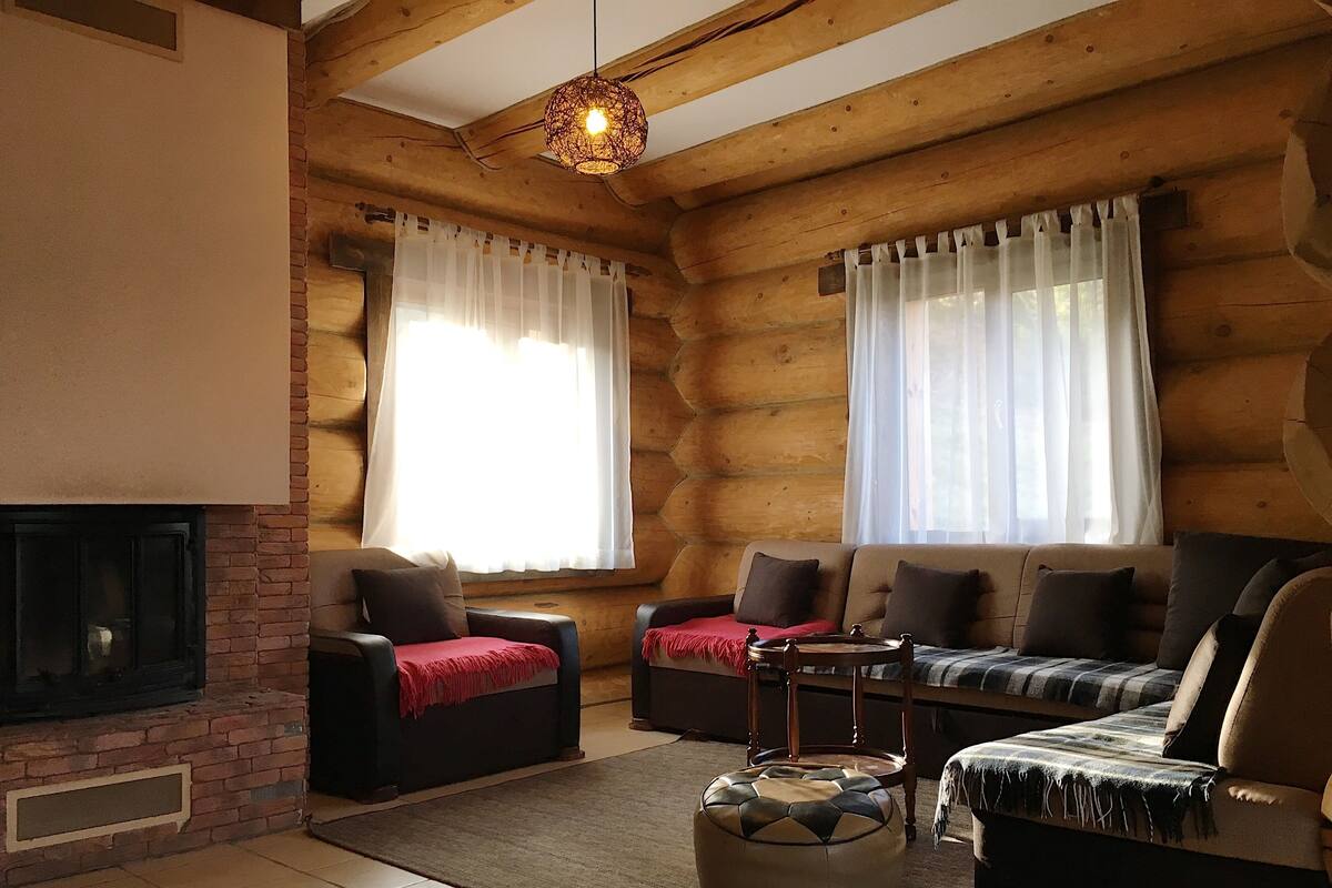 A log cabin interior with a fireplace, two sofas, a coffee table, and a patterned ottoman.