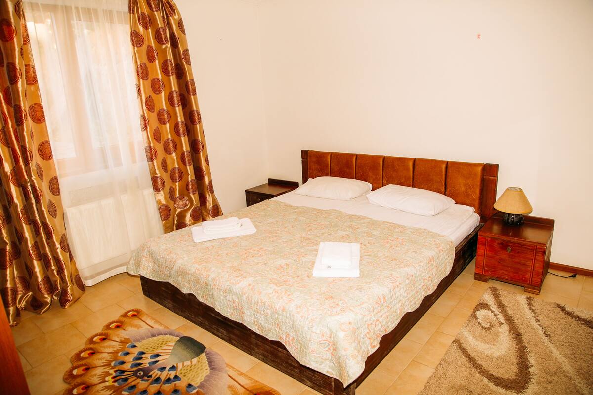 A bedroom with a large bed, a wooden headboard, a nightstand with a lamp, and a peacock-patterned rug.