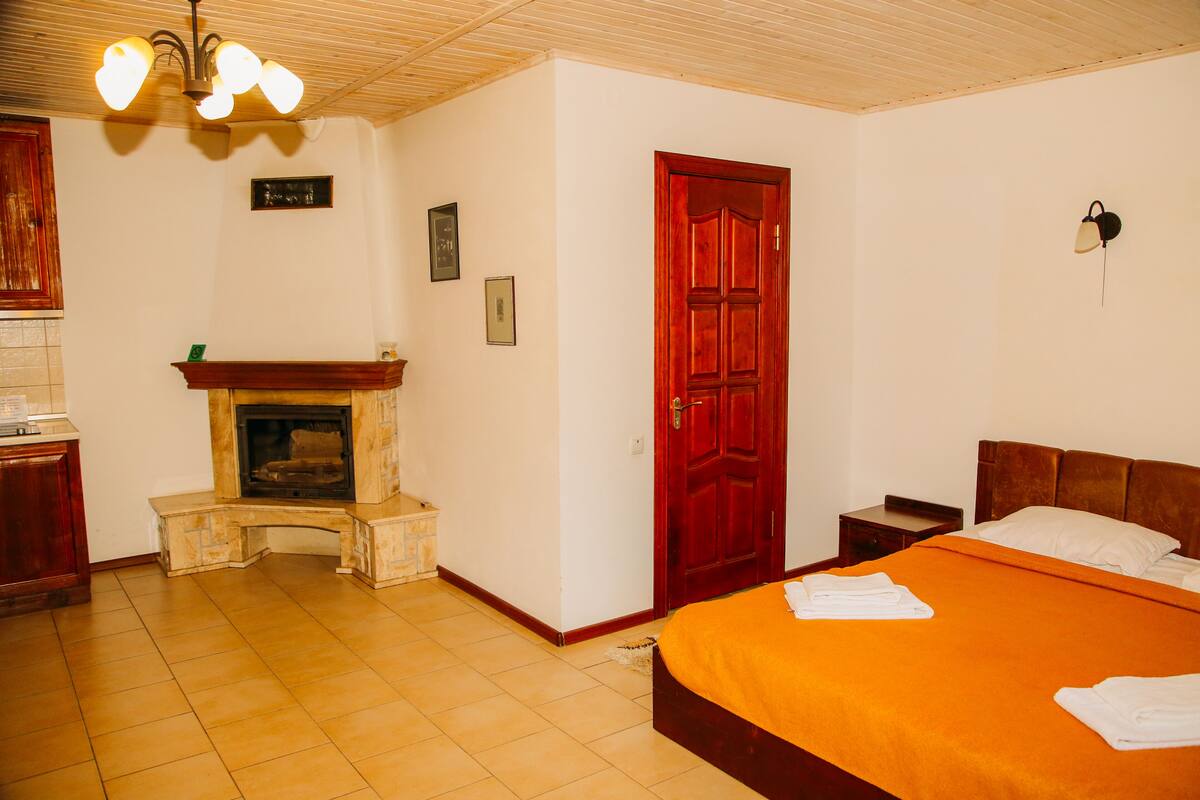 A hotel room with a fireplace, a bed with an orange bedspread, a wooden headboard, a red door, and a wall-mounted light fixture.