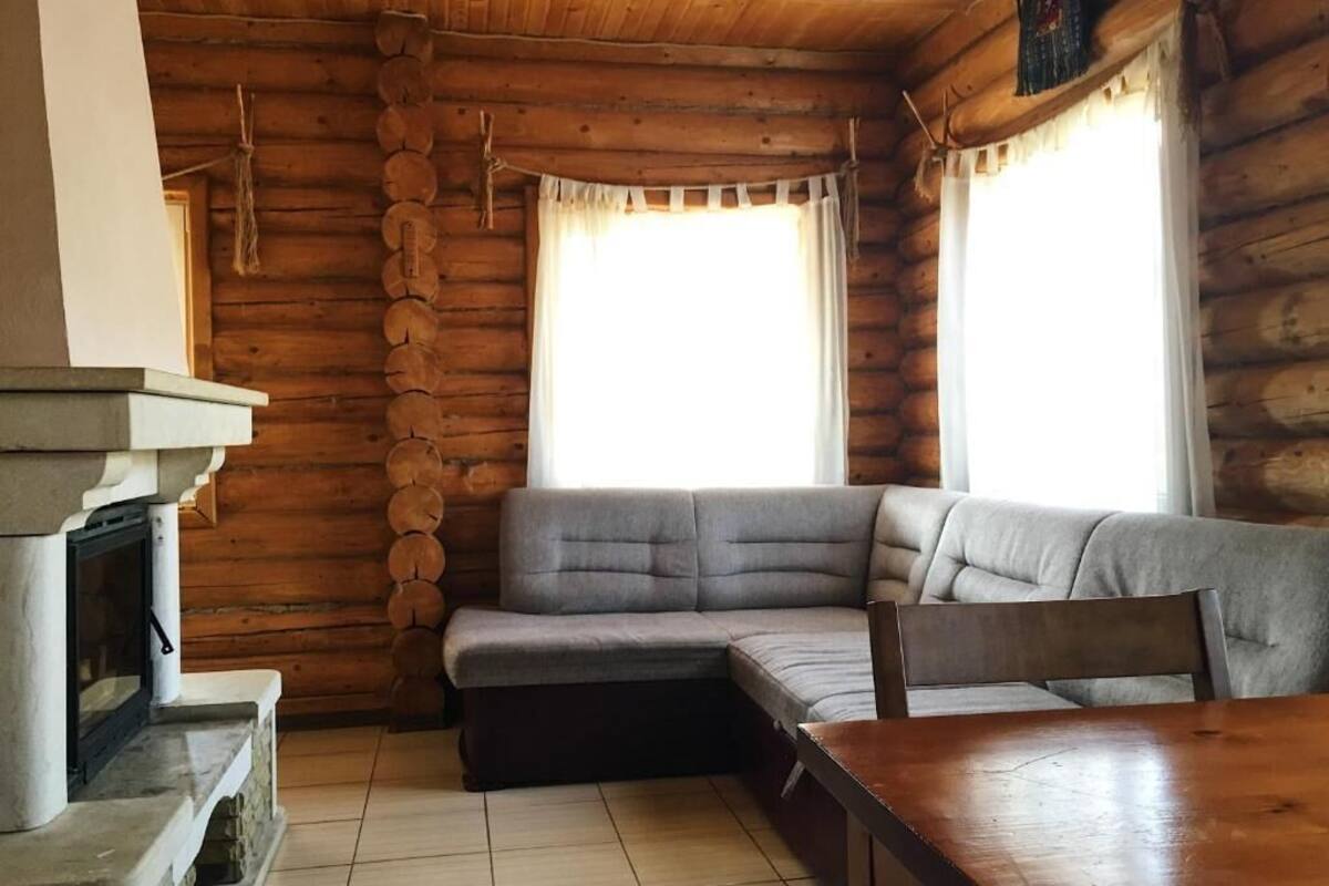 A log cabin interior with a stone fireplace, a gray sectional sofa, and a wooden dining table.