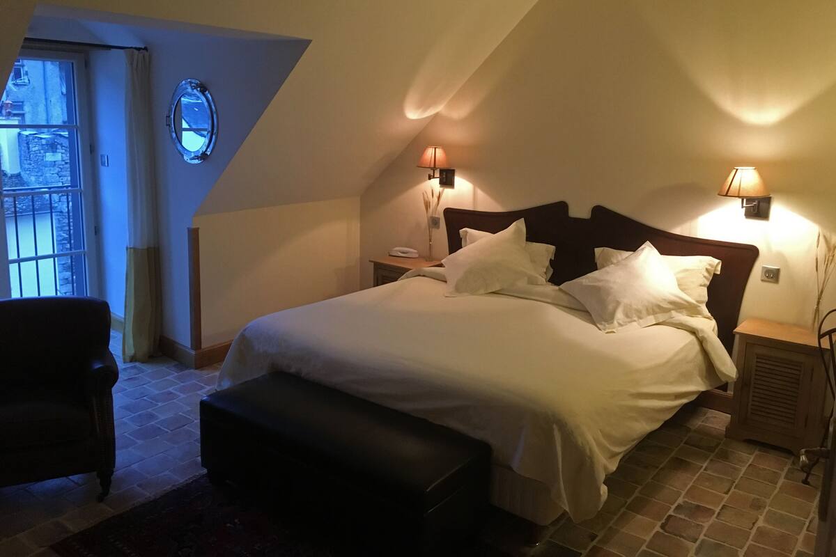 A bedroom with a large bed, two bedside lamps, a small table, a chair, and a window with a view of the outside.