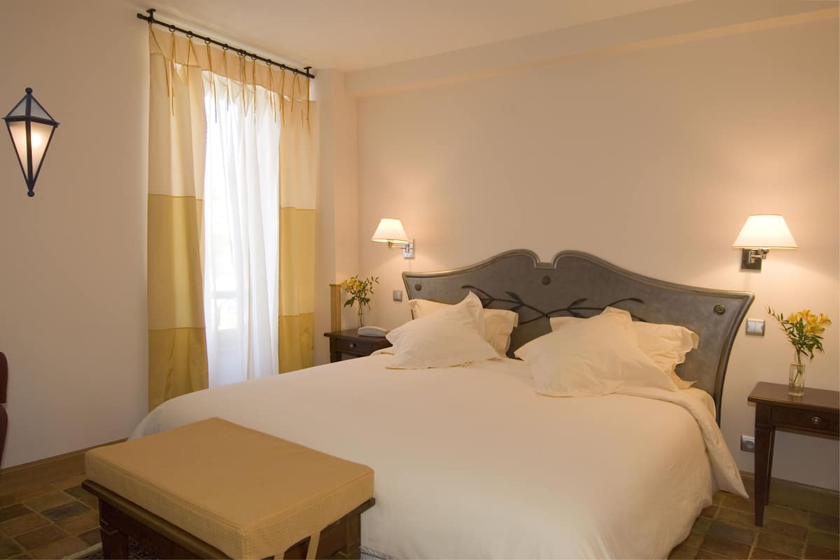 A neatly made bed with white linens, a bedside table with a lamp, and a window with curtains.