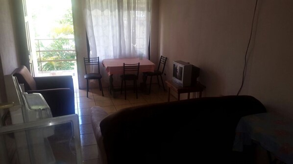 Una sala de estar con sofá, televisor en un mueble bajo, una mesa de comedor con sillas y una ventana con cortinas.