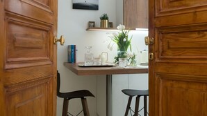 Una puerta de madera con herrajes de latón, una pequeña mesa con un decantador de vidrio, dos taburetes negros, un jarrón con flores blancas y un cuadro en la pared.