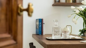 Una mesa de madera con una bandeja que contiene un decantador de vidrio, una copa y una servilleta blanca. En el estante superior hay libros titulados 'ROM' y 'COURTESY HOUSE OF ENGLAND'.
