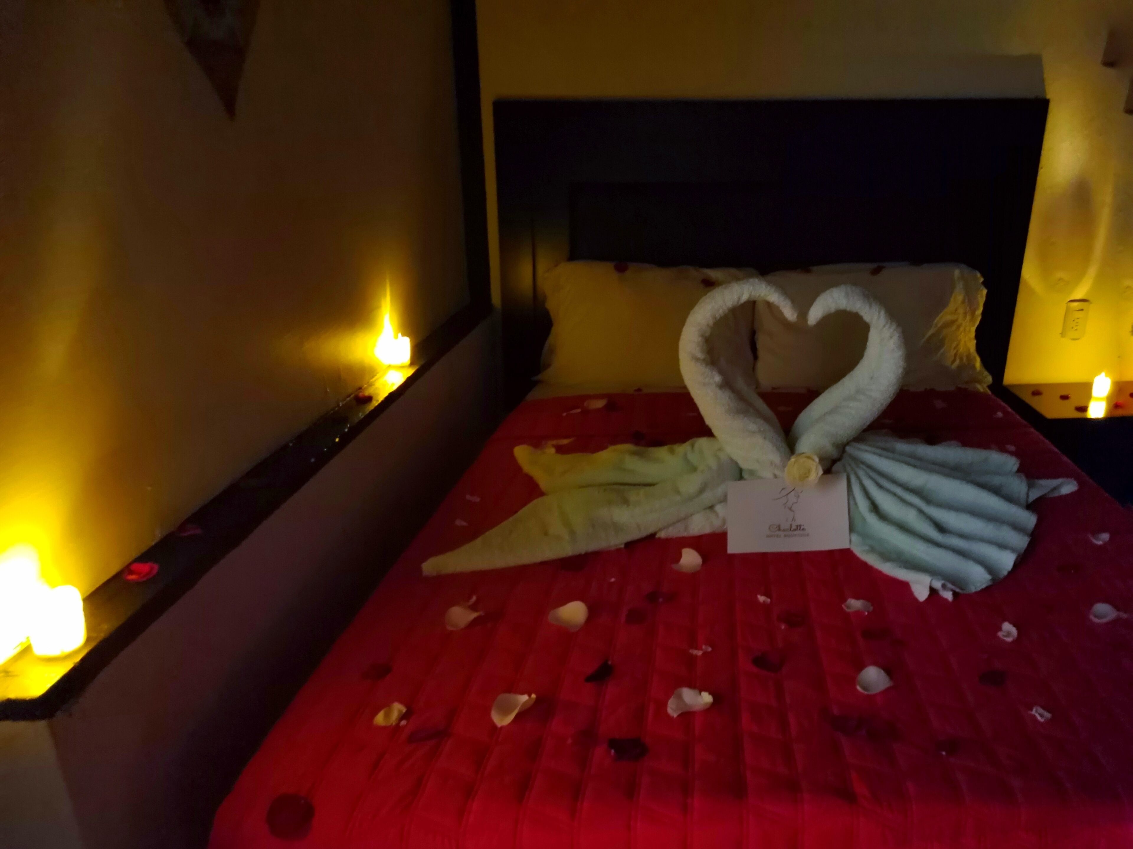A bed with a red bedspread, a heart-shaped towel arrangement, and candles on the nightstands.