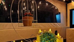 A room with a wooden ceiling, a window with a view, a table with candles and a basket of greenery, and a hanging plant in a wire cage.