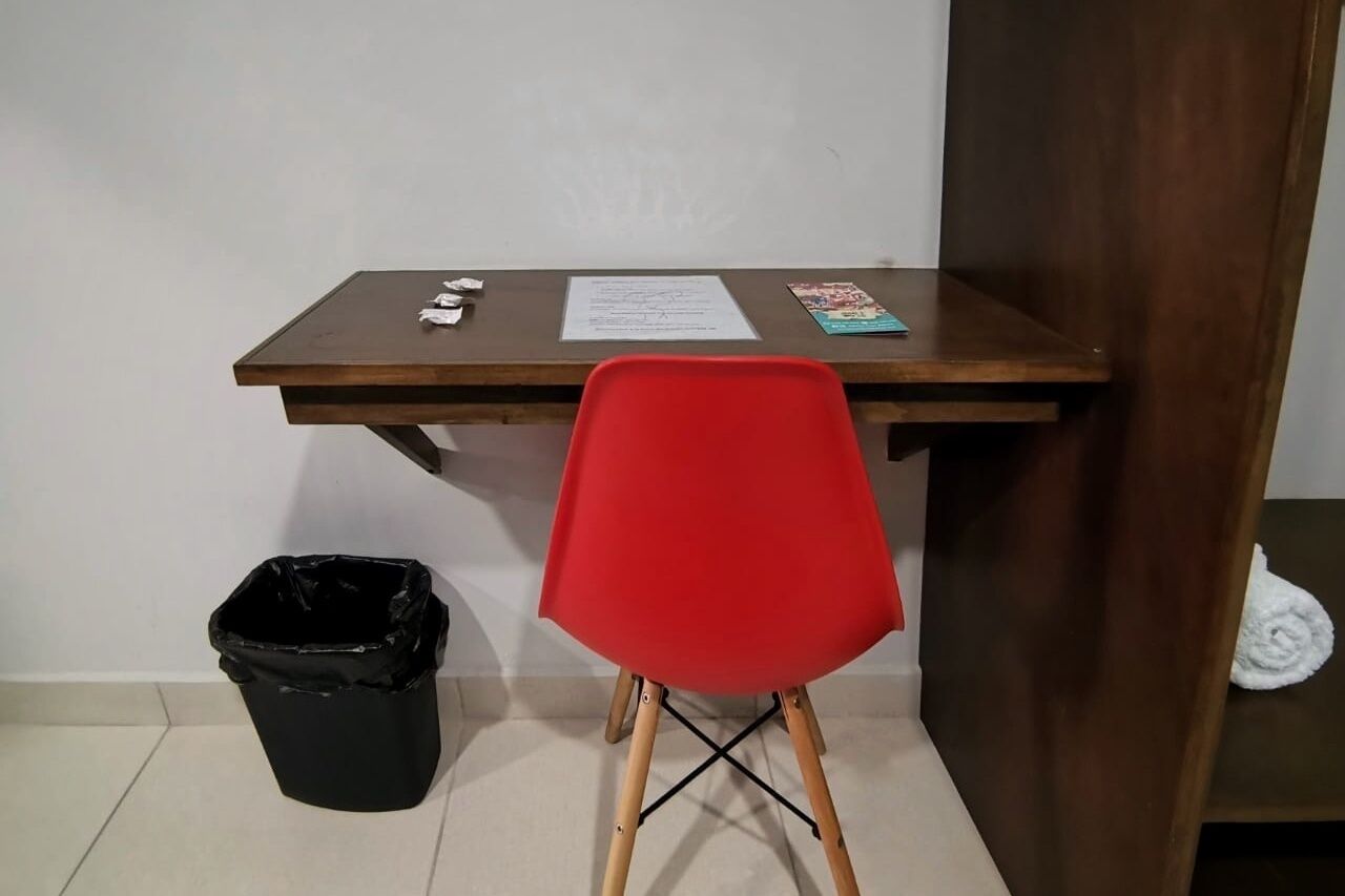 A small room with a wooden desk, a red chair, a black trash bin, and a white towel on a shelf.