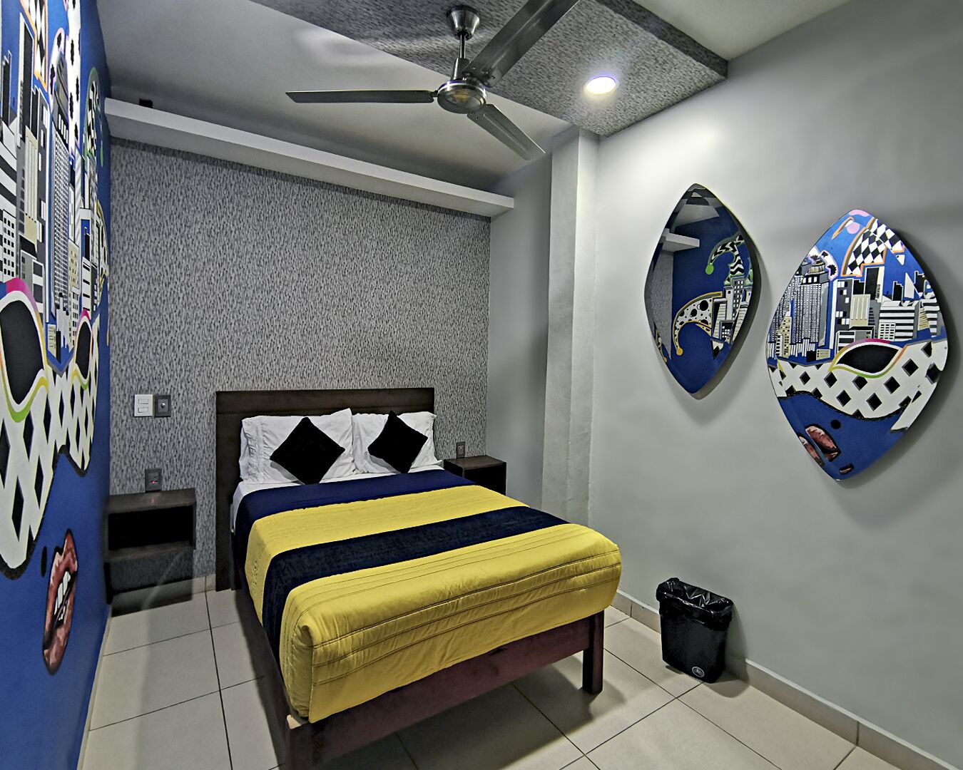 Un dormitorio con una cama, un ventilador de techo, una pared con un mural de paisaje urbano y un cubo de basura.