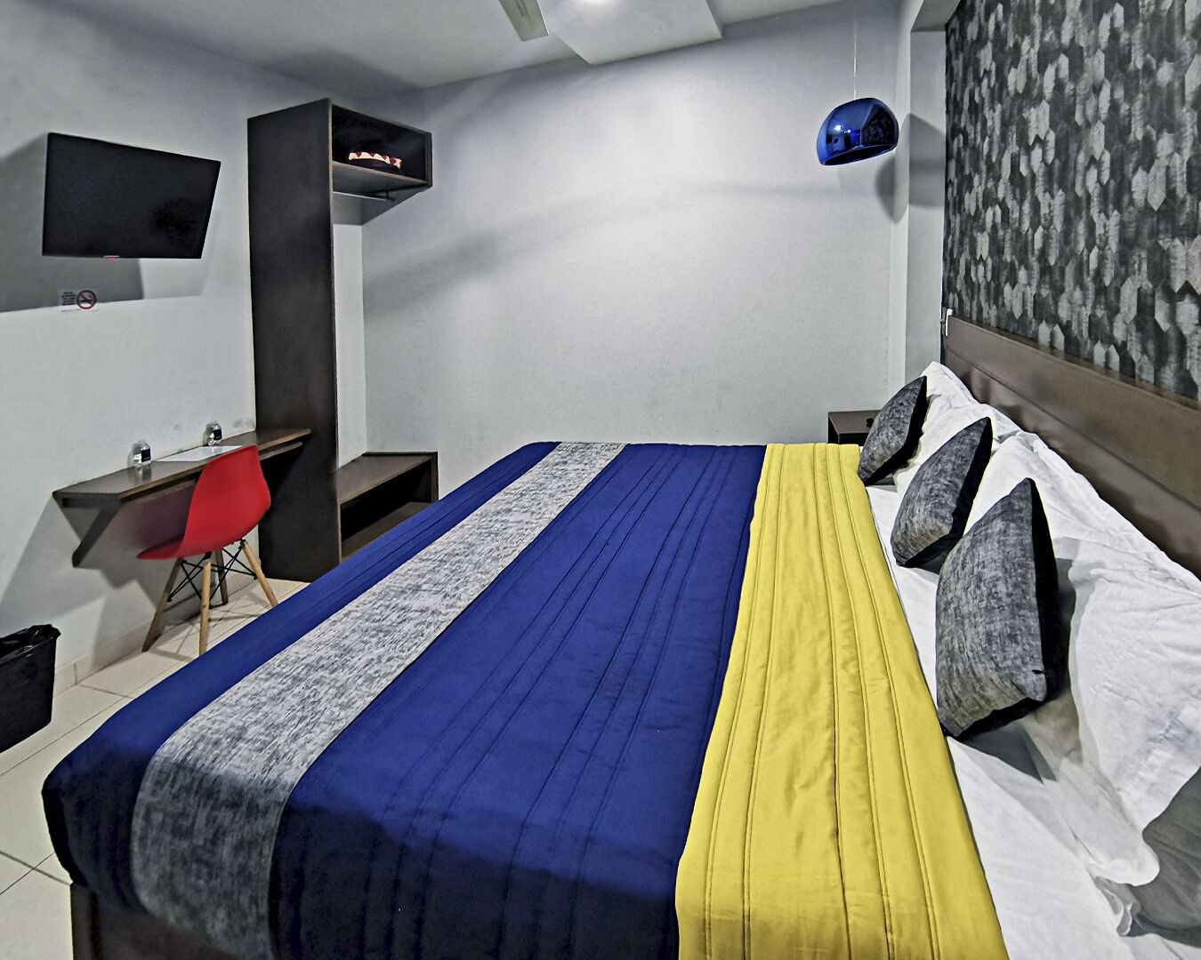 Une chambre à coucher comprenant un lit, un bureau, une chaise, un téléviseur et une étagère fixée au mur.