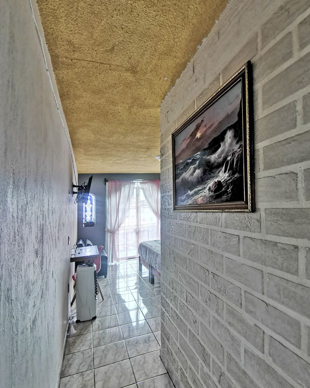 Un pasillo estrecho con suelo de baldosas, pared de ladrillo y un cuadro enmarcado que representa un mar embravecido.