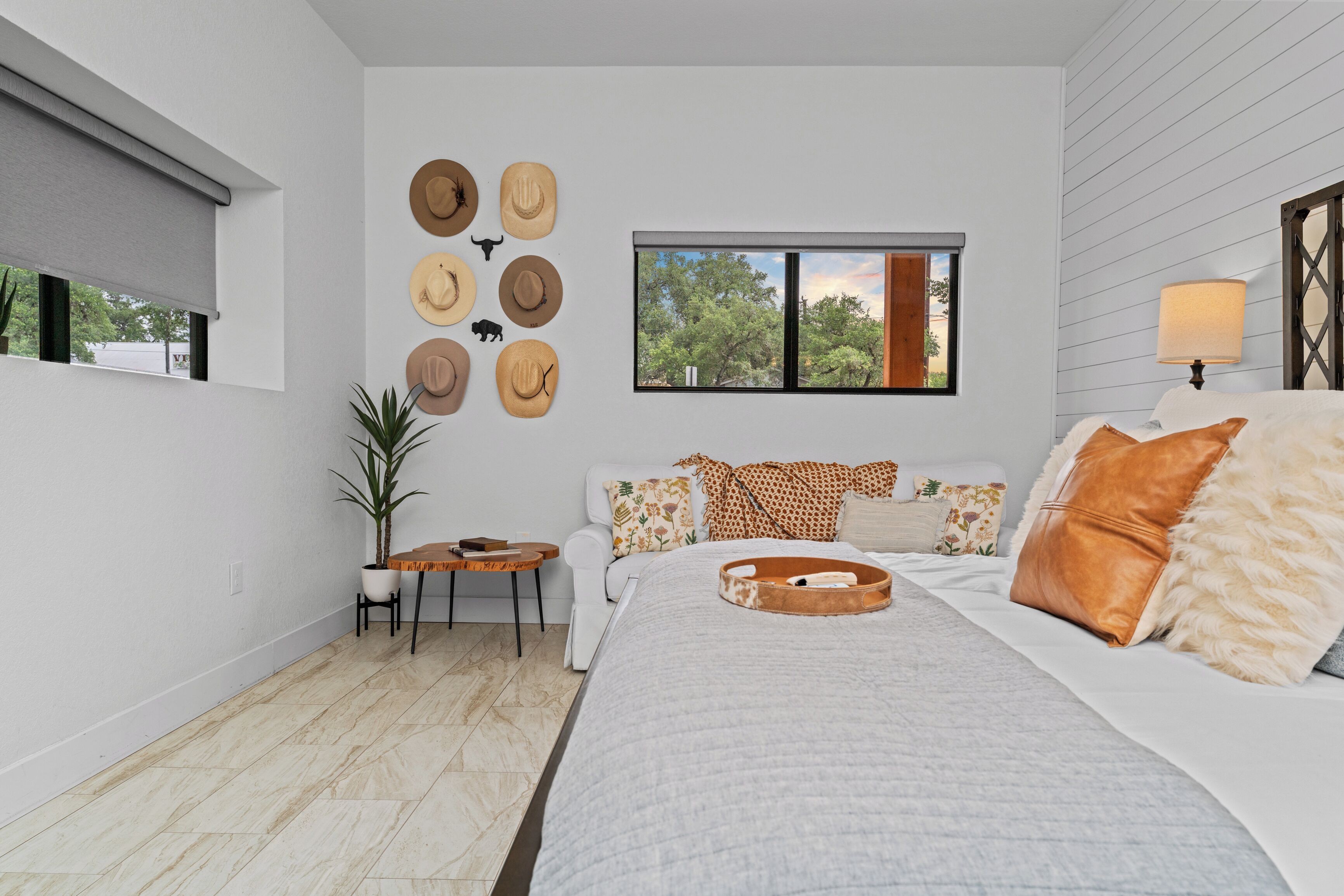 A bedroom with a large bed, a small table with a plant, a window with a view of trees, and a wall with hats.