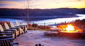 Un patio de piedra con sillas Adirondack y una chimenea, con vistas a un lago al atardecer.