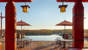 Un patio al aire libre con dos mesas y sillas, cada una bajo un paraguas a rayas, con vistas a un lago y un paisaje boscoso de fondo.