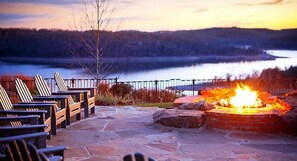 A stone patio with Adirondack chairs and a fire pit overlooking a lake at sunset.