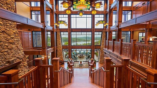 Un interior espacioso con una escalera central, barandillas de madera y grandes ventanales que ofrecen vistas al exterior.