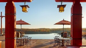 Un patio al aire libre con dos mesas y sillas, cada una bajo un paraguas a rayas, con vistas a un lago y un paisaje boscoso de fondo.