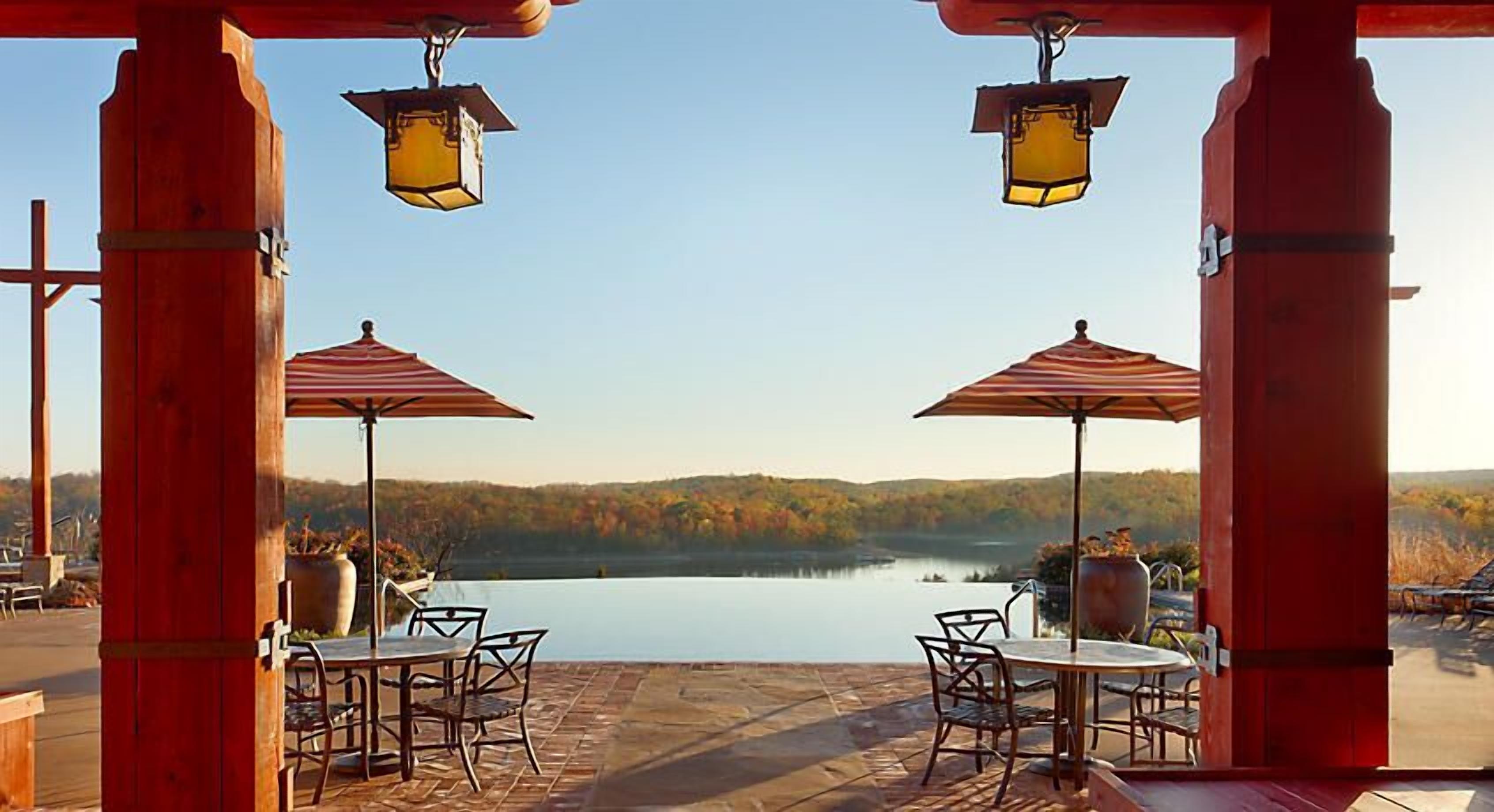 An outdoor patio with two tables and chairs, each under a striped umbrella, overlooking a lake with a forested backdrop.
