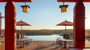 An outdoor patio with two tables and chairs, each under a striped umbrella, overlooking a lake with a forested backdrop.