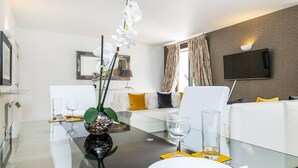 A modern living room with a glass dining table, white sofa, and a flat-screen TV mounted on the wall. - Elegant Two bedroom in the heart of Kensington (London)