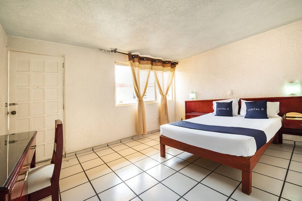 A hotel room with a bed, a desk, a chair, a window with curtains, and a nightstand with a lamp.