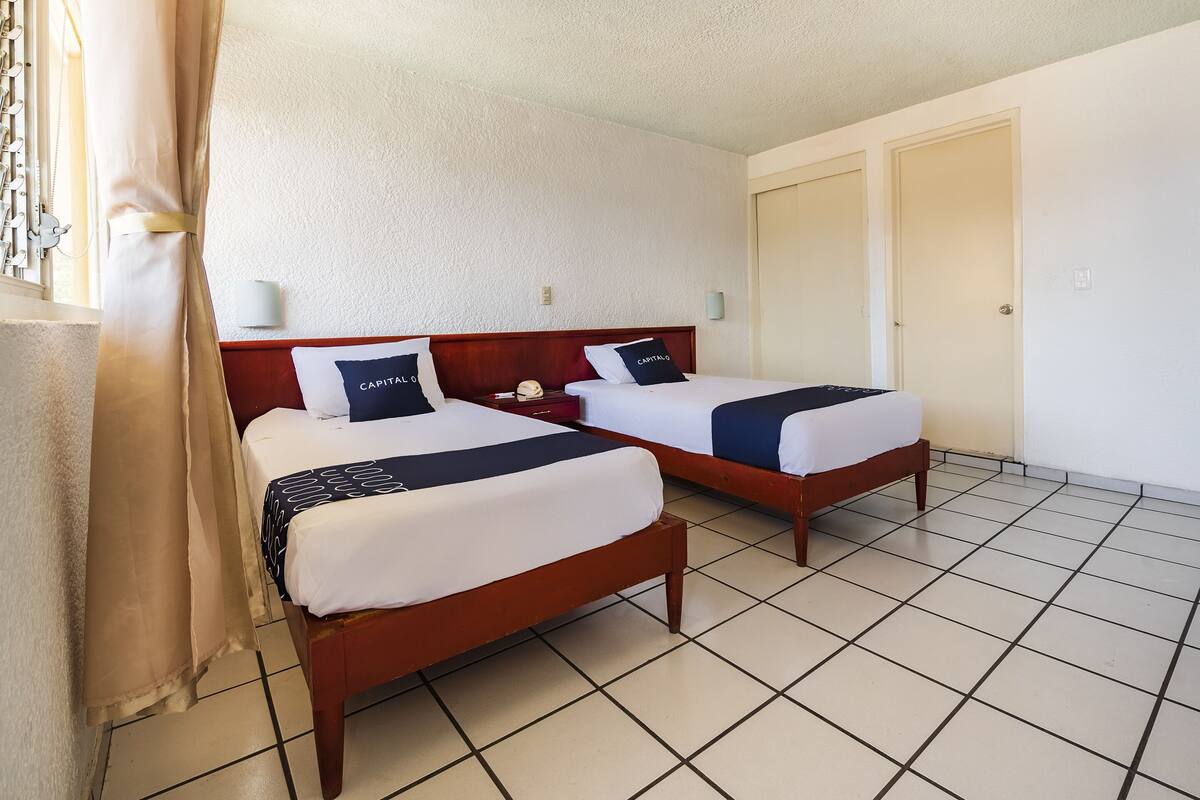 A hotel room with two beds, a tiled floor, and a plain white wall.